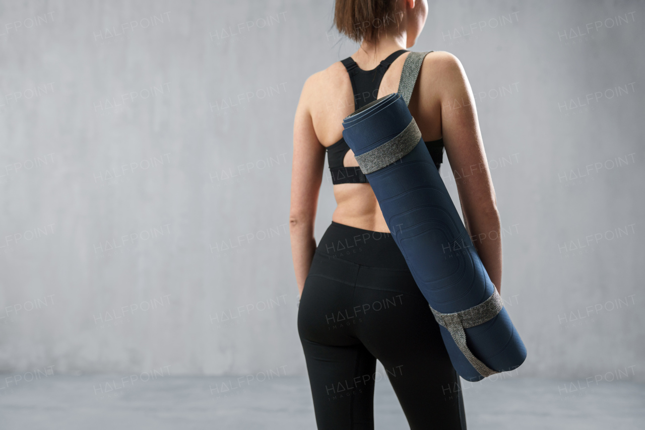 A rear view of sports woman in fashion black sport clothes carrying fitness mat in gym, over gray background