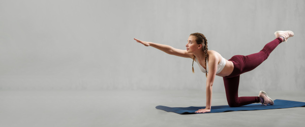 A sports woman in fashion sport clothes working out against grey wall, doing yoga, pilates balancing exercise.