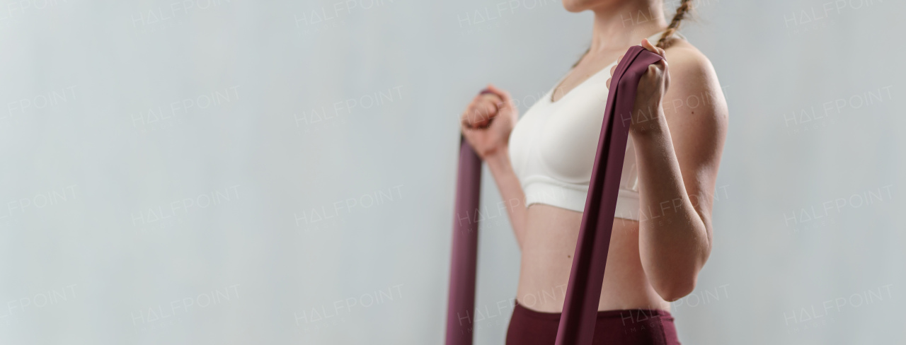 A close-up of sports woman in fashion sportswear exercising with elastic band stretch over greay background.