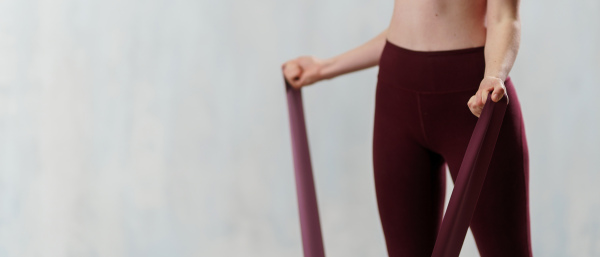 A close-up of sports woman in fashion sportswear exercising with elastic band stretch over greay background.