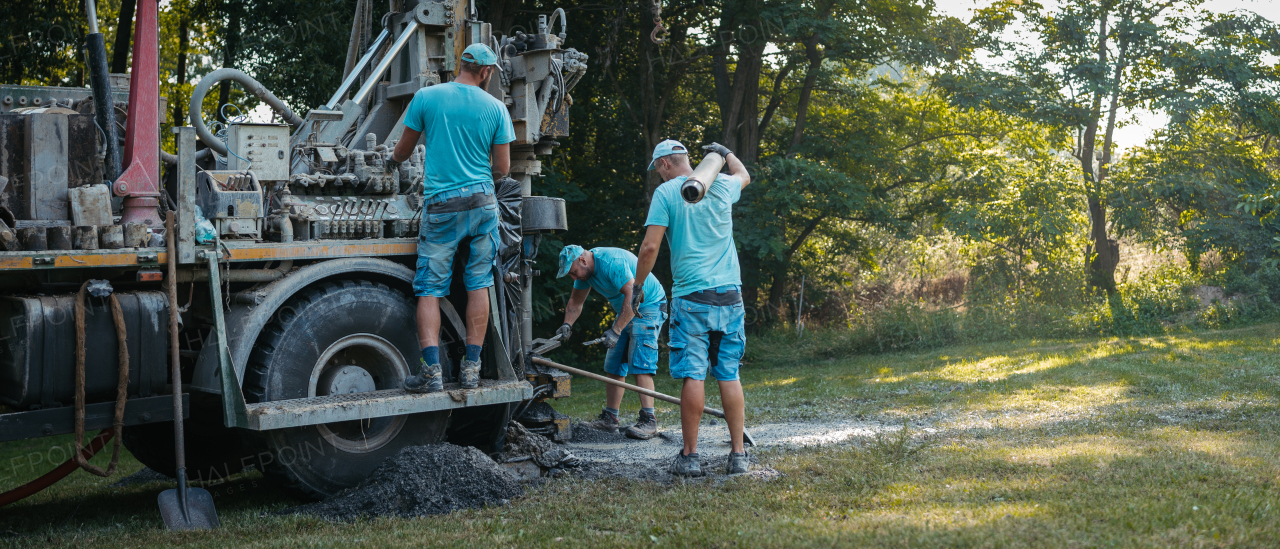 Water well drilling rig, truck preparing to boring dowin into the earth. Professional workers operating drilling machine, shoveling rubble. A rotary drill rig using bits to bore into ground and loosing the soil and rocks. Mud and muddy water is spitting out when drilling borewell. Banner with copy space.