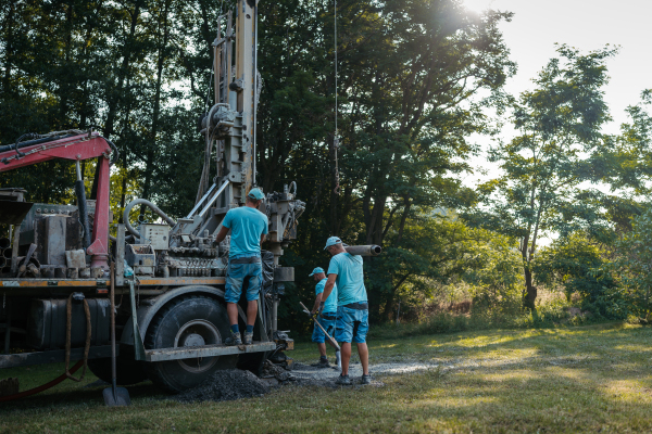 Water well drilling rig, truck preparing to boring dowin into the earth. Professional workers operating drilling machine, shoveling rubble. A rotary drill rig using bits to bore into ground and loosing the soil and rocks. Mud and muddy water is spitting out when drilling borewell. Banner with copy space.