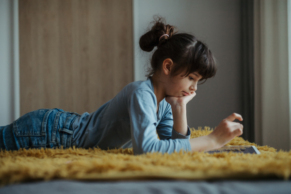 Cute girl lying on belly on the floor, scrolling on smartphone. Using social media. Side view.