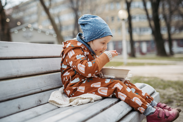 Cute toddler girl eating lunch in park, sitting on bench. Picnic in city park during cold spring day.