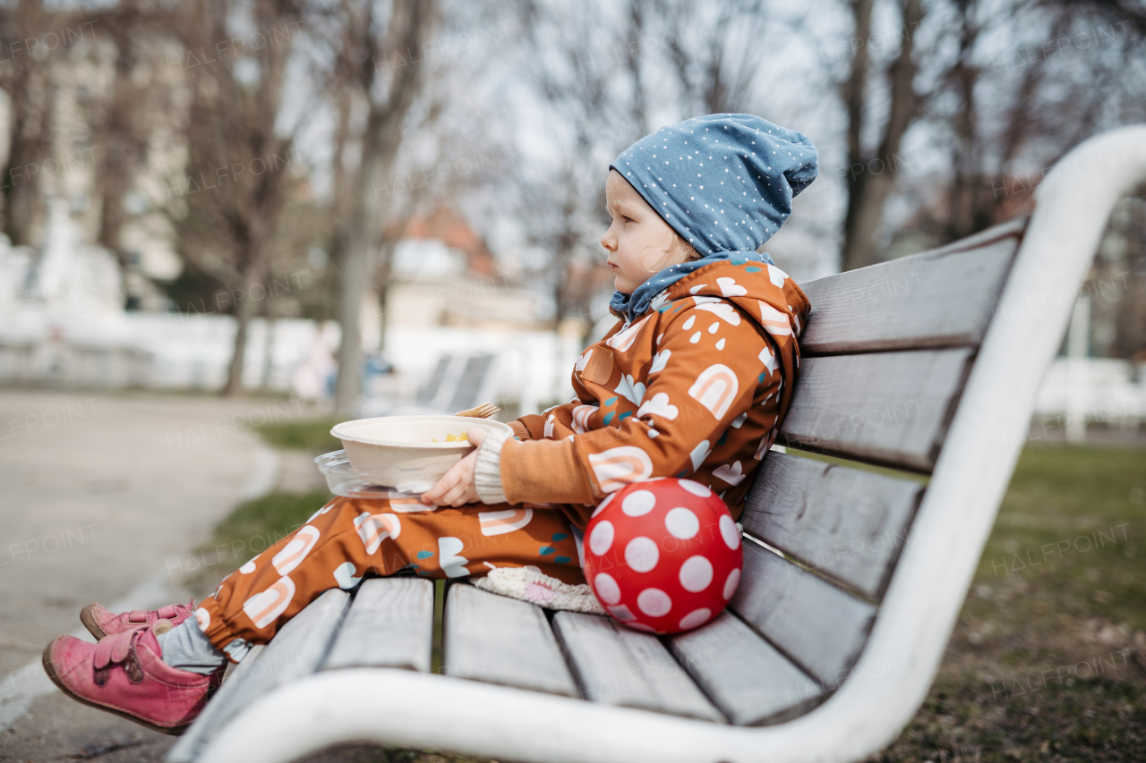 Cute toddler girl eating lunch in park, sitting on bench. Picnic in city park during cold spring day.