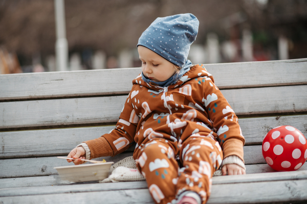 Cute toddler girl eating lunch in park, sitting on bench. Picnic in city park during cold spring day.