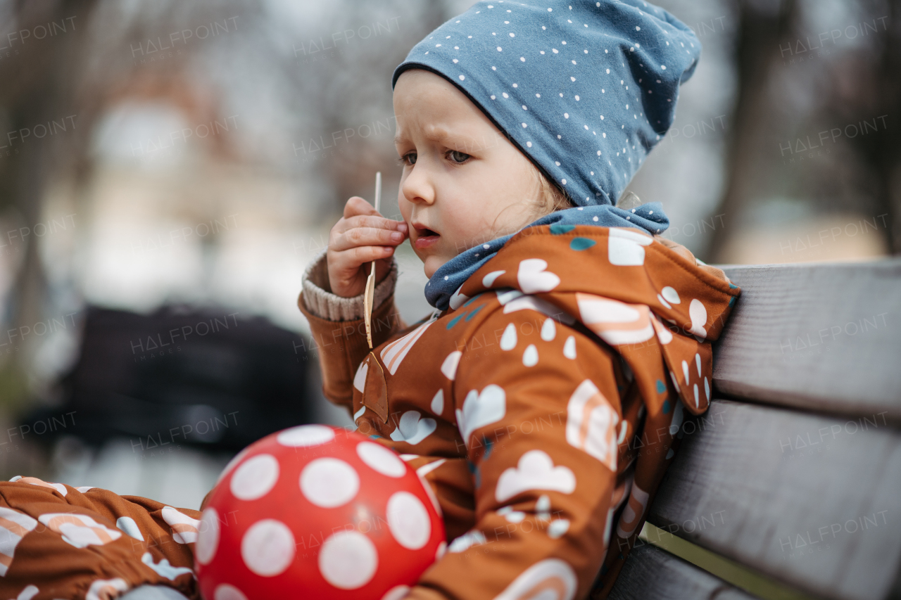 Cute toddler girl eating lunch in park, sitting on bench. Picnic in city park during cold spring day.