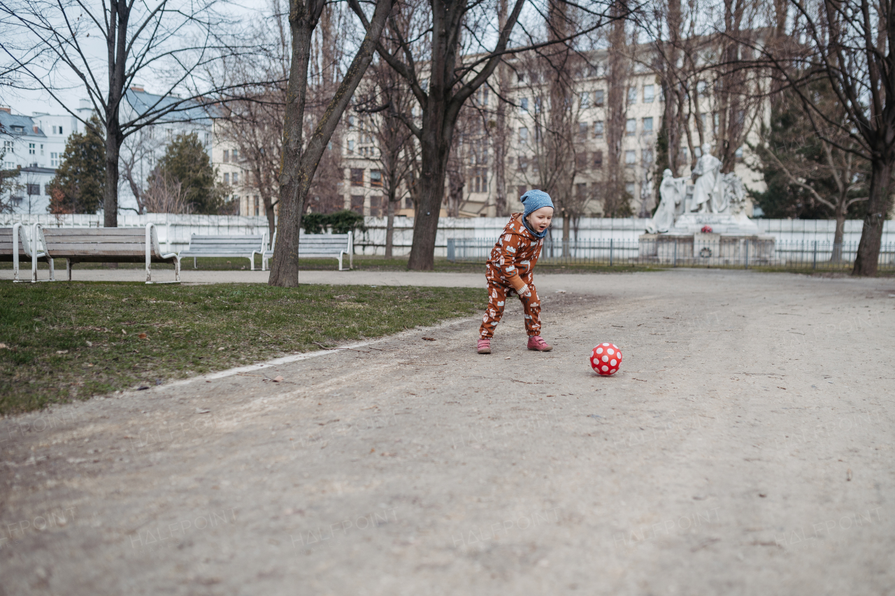 Cute toddler girl playing outdoor in playground with ball. Girl in softshell bodysuit spending time in city park.