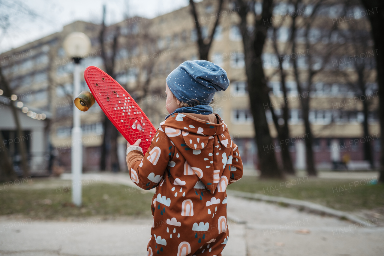 Cute toddler girl playing outdoor in playground with skateboard. Girl in softshell bodysuit spending time in city park.