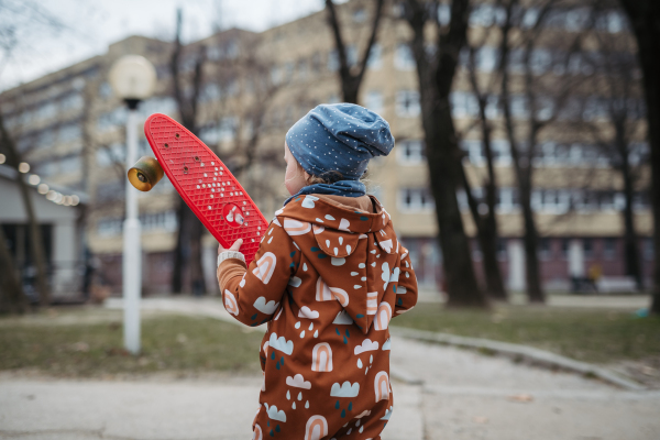 Cute toddler girl playing outdoor in playground with skateboard. Girl in softshell bodysuit spending time in city park.