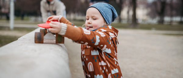 Cute toddler girl playing outdoor in playground with skateboard. Girl in softshell bodysuit spending time in city park.