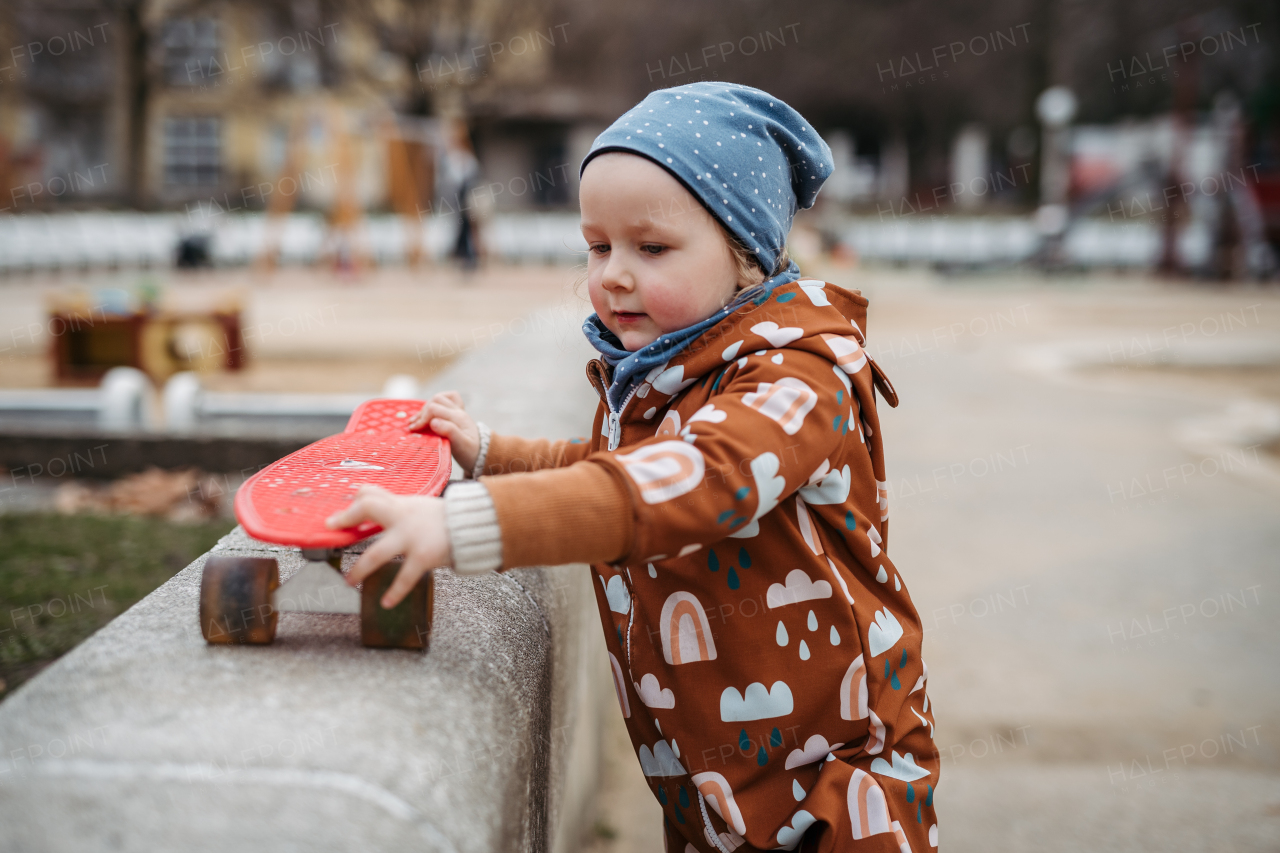 Cute toddler girl playing outdoor in playground with skateboard. Girl in softshell bodysuit spending time in city park.