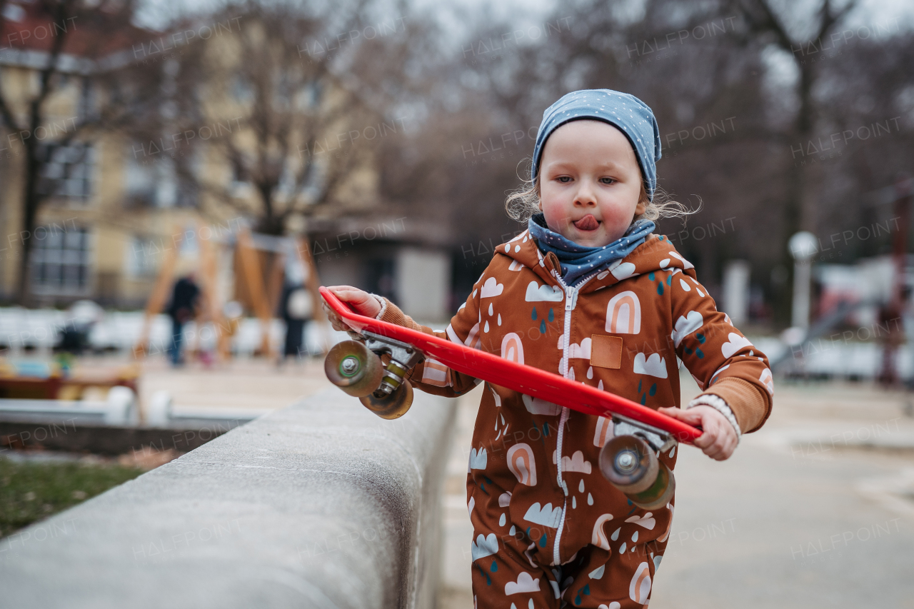 Cute toddler girl playing outdoor in playground with skateboard. Girl in softshell bodysuit spending time in city park.