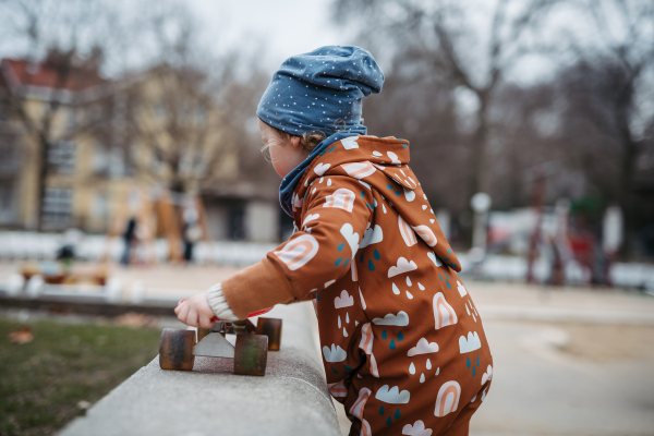 Cute toddler girl playing outdoor in playground with skateboard. Girl in softshell bodysuit spending time in city park.