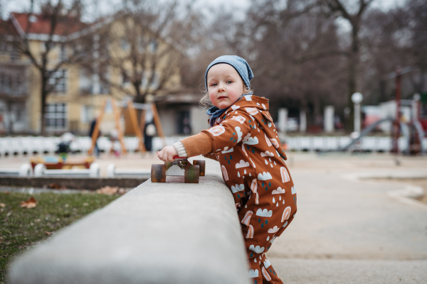 Cute toddler girl playing outdoor in playground with skateboard. Girl in softshell bodysuit spending time in city park.