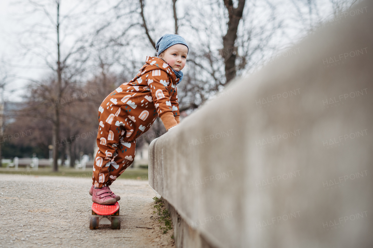 Cute toddler girl playing outdoor in playground with skateboard. Girl in softshell bodysuit spending time in city park.