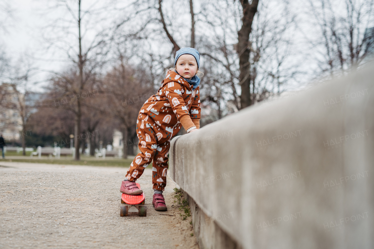 Cute toddler girl playing outdoor in playground with skateboard. Girl in softshell bodysuit spending time in city park.