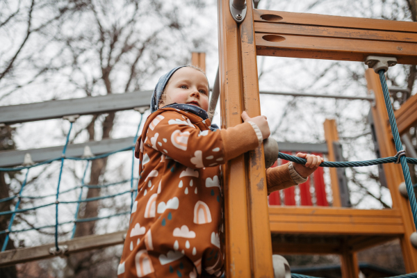 Cute toddler girl playing outdoor in playground, climbing jungle gym. Girl in softshell bodysuit spending time in city park.