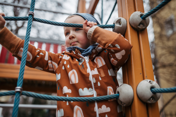 Cute toddler girl playing outdoor in playground, climbing jungle gym. Girl in softshell bodysuit spending time in city park.