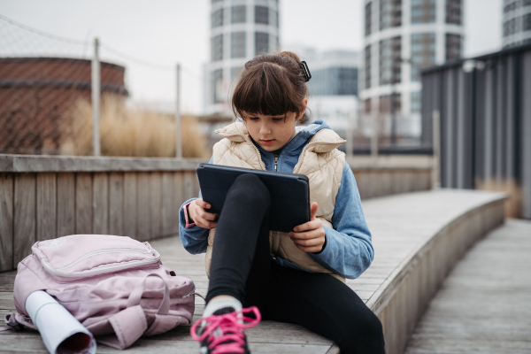 Girl sitting in front of school building, scrolling on tablet. Using social media. Online bullying or cyberbullying for a young schoolgirl.