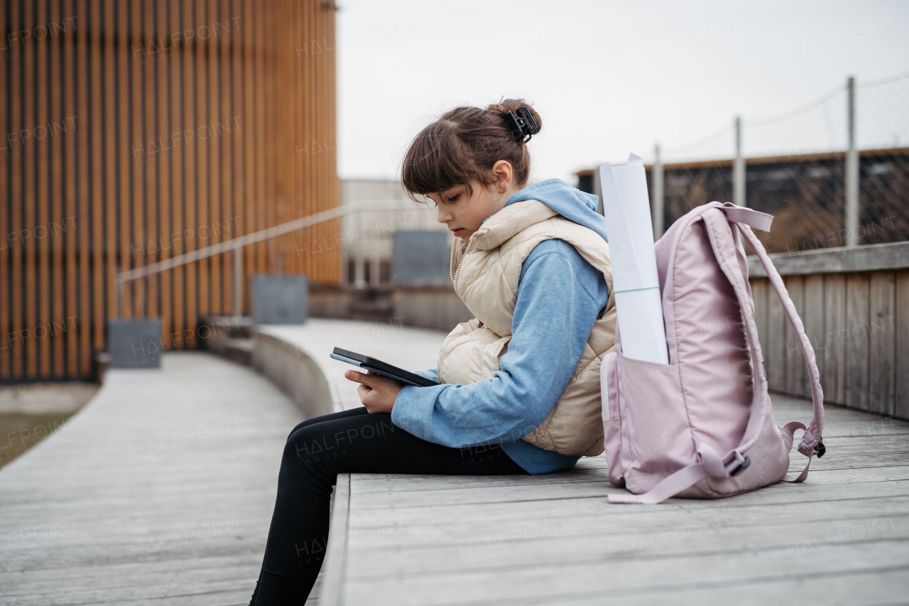 Girl sitting in front of school building, scrolling on tablet. Using social media. Online bullying or cyberbullying for a young schoolgirl.