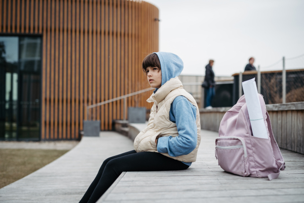 Sad girl alone, sitting in front of school building, waiting for parents. Young schoolgirl with no friends, feeling lonely. Mental health problems.