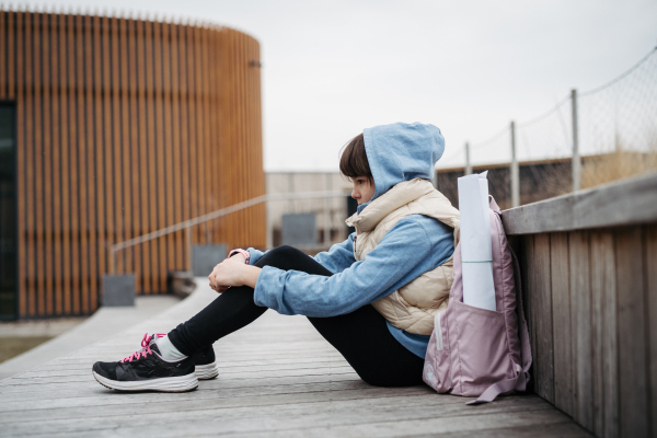 Sad girl alone, sitting in front of school building, waiting for parents. Young schoolgirl with no friends, feeling lonely. Mental health problems.