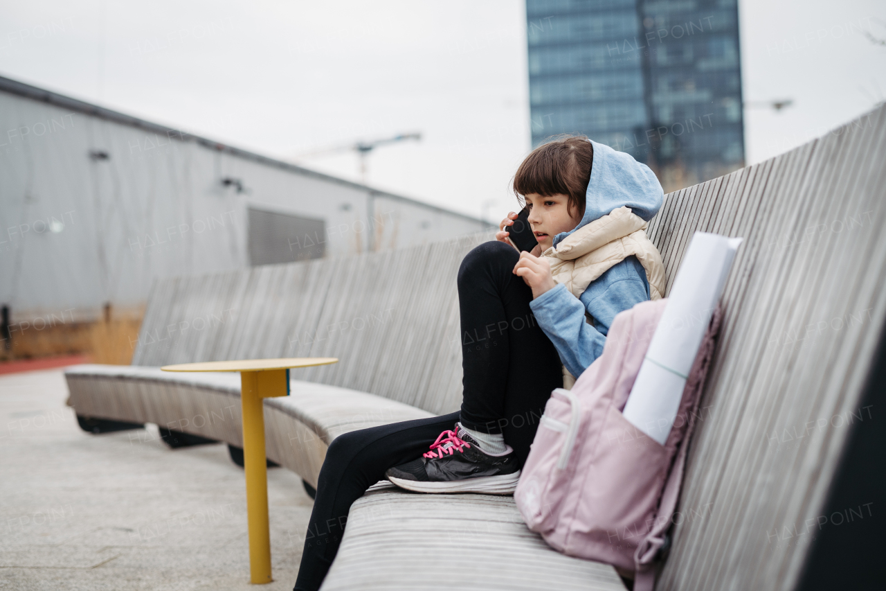 Sad girl phone calling, sitting in front of school building, waiting for parents. Young schoolgirl with no friends, feeling lonely. Mental health problems.