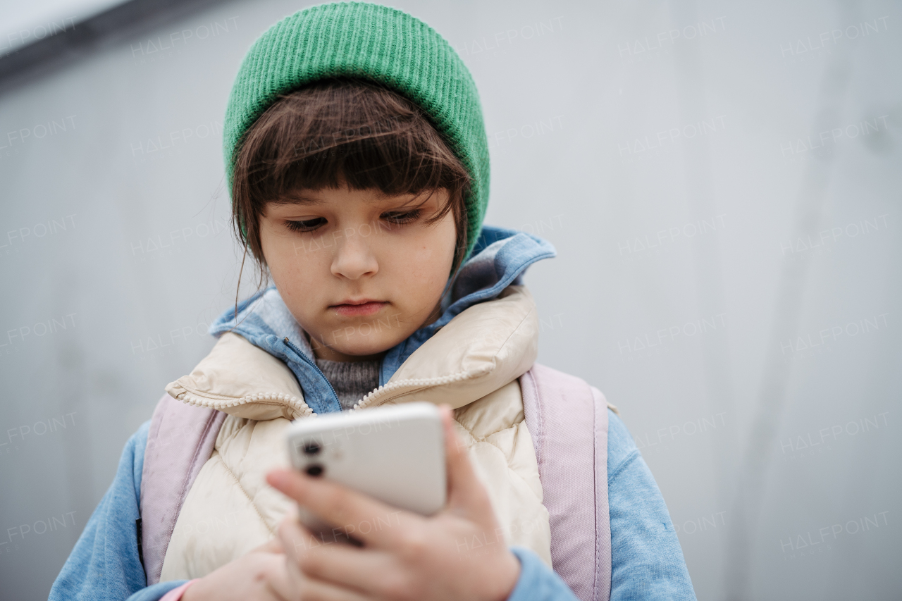 Girl in front of school building, scrolling on smartphone. Using social media. Online bullying or cyberbullying for a young schoolgirl.