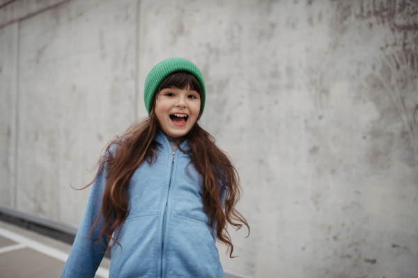 Outdoor portrait of cute young girl with beanie hat, laughing. Girl with a long hair standing on playground in the city.