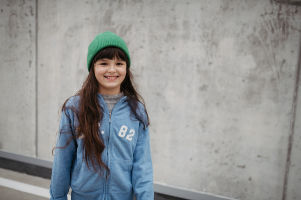 Outdoor portrait of cute young girl with beanie hat, laughing. Girl with a long hair standing on playground in the city.