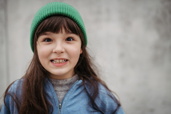 Outdoor portrait of cute young girl with beanie hat, laughing. Girl with a long hair standing on playground in the city.