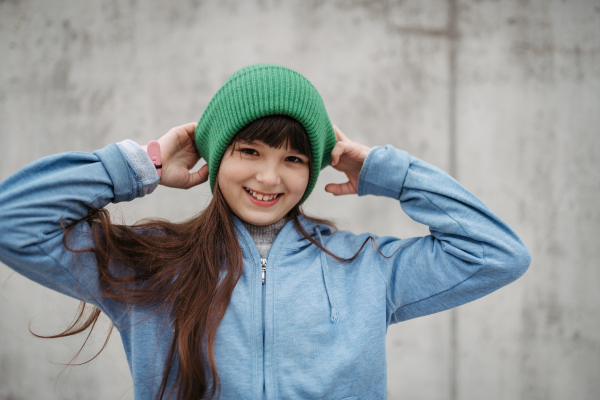 Outdoor portrait of cute young girl with beanie hat, laughing. Girl with a long hair standing on playground in the city.