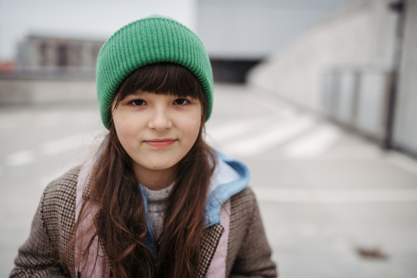 Outdoor portrait of cute young girl with beanie hat, laughing. Girl with a long hair standing on playground in the city.