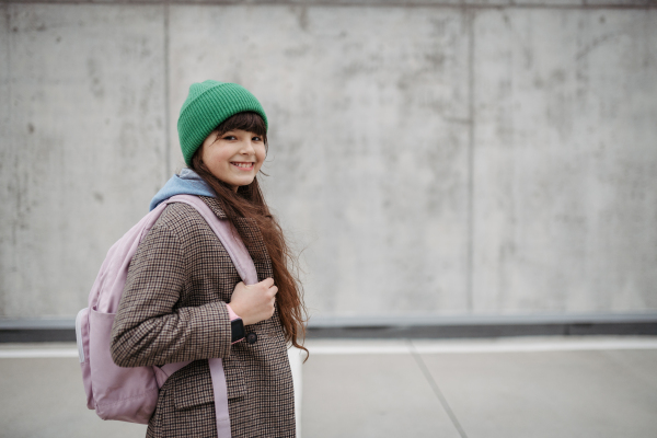 Outdoor portrait of cute young girl with beanie hat, laughing. Girl with a long hair standing on playground in the city.