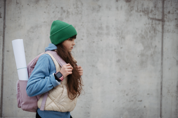 Outdoor portrait of sad young girl with beanie hat and backpack. Girl with long hair going to school, feeling anxious, depressed.