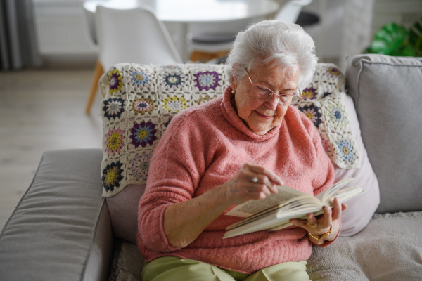 Beautiful elderly woman reading book at home, relaxing moment, hygge for senior. Grandmother with reading glasses and captivating book.