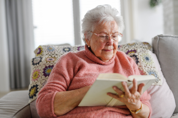 Beautiful elderly woman reading book at home, relaxing moment, hygge for senior. Grandmother with reading glasses and captivating book.