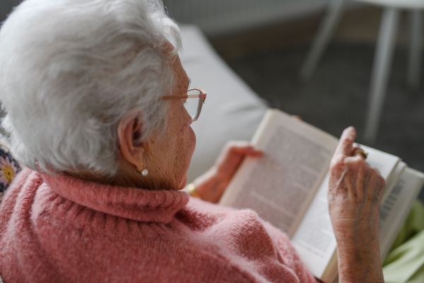 Beautiful elderly woman reading book at home, relaxing moment, hygge for senior. Grandmother with reading glasses and captivating book.