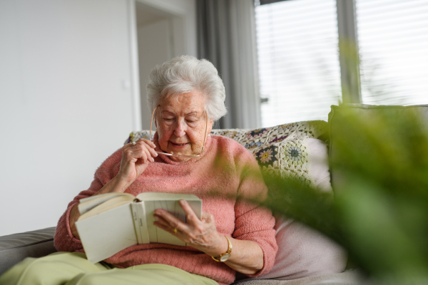 Beautiful elderly woman reading book at home, relaxing moment, hygge for senior. Grandmother with reading glasses and captivating book.