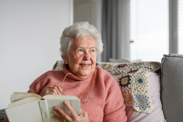 Beautiful elderly woman reading book at home, relaxing moment, hygge for senior. Grandmother with reading glasses and captivating book.