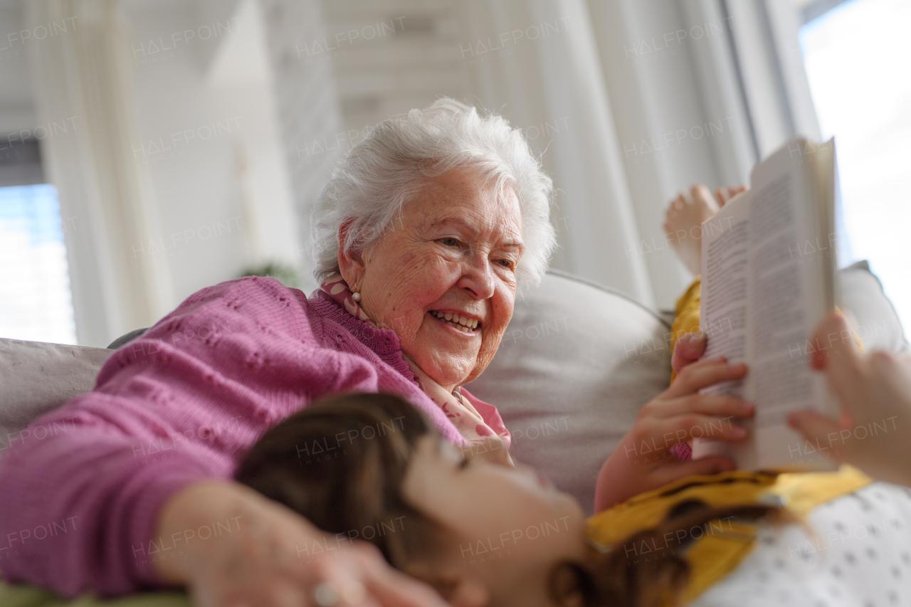 Cute girl lying head on gradmother knees and reading book, grandma caressing her hair. Portrait of an elderly woman spending time with granddaughter.