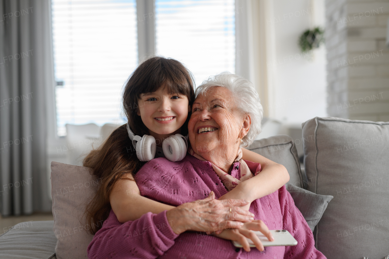 Cute girl hugging gradmother from behind, looking at camera and smiling. Portrait of an elderly woman spending time with granddaughter.