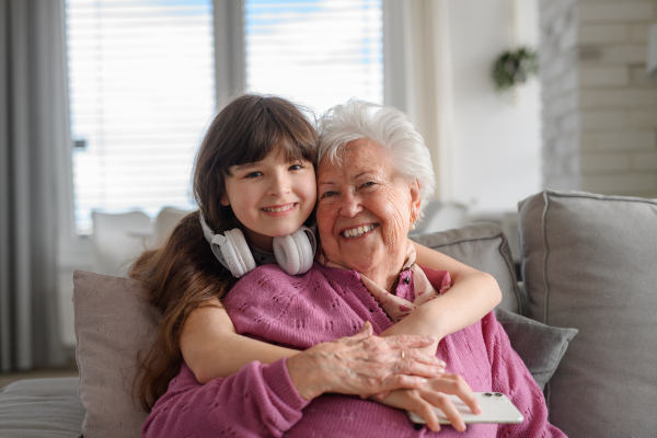 Cute girl hugging gradmother from behind, looking at camera and smiling. Portrait of an elderly woman spending time with granddaughter.