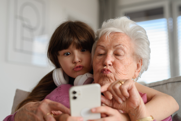 Grandmother with cute girl taking selfie with smartphone, making funny face. Portrait of an elderly woman spending time with a granddaughter.