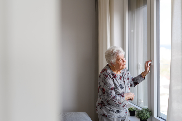 Senior woman looking out of window, spending time alone in apartment. Concept of loneliness and dependence of retired people.