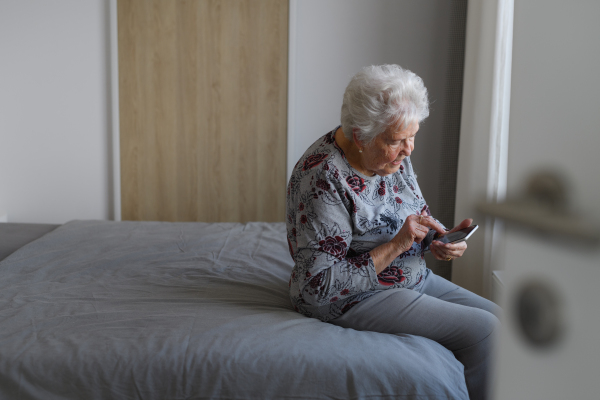 Senior woman at home, texting with friends or family member, sitting on bed. Older woman living alone, enjoying peaceful weekend day.