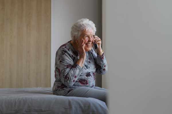 Senior woman at home, phone calling with family member, sitting on bed. Older woman living alone, enjoying peaceful weekend day.