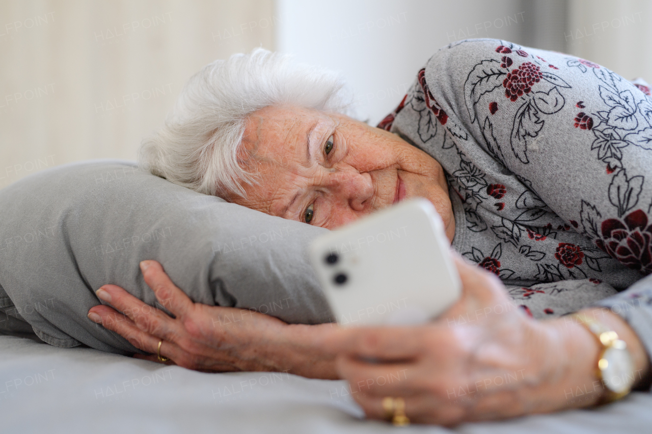 Senior woman lying in bed, looking at smartphone and smiling. Older woman using modern technology, digital skill and literacy.