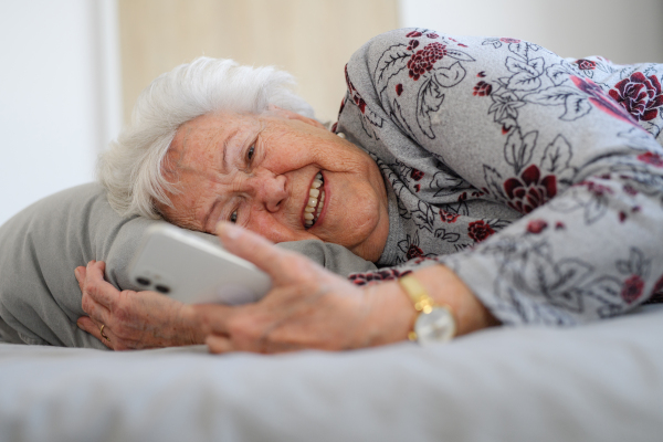 Senior woman lying in bed, looking at smartphone and smiling. Older woman using modern technology, digital skill and literacy.